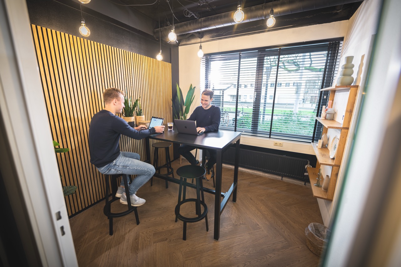 Twee mannen werken op laptops aan een hoge tafel in een modern ingerichte kantoorruimte met houten vloer en lamellenwand.