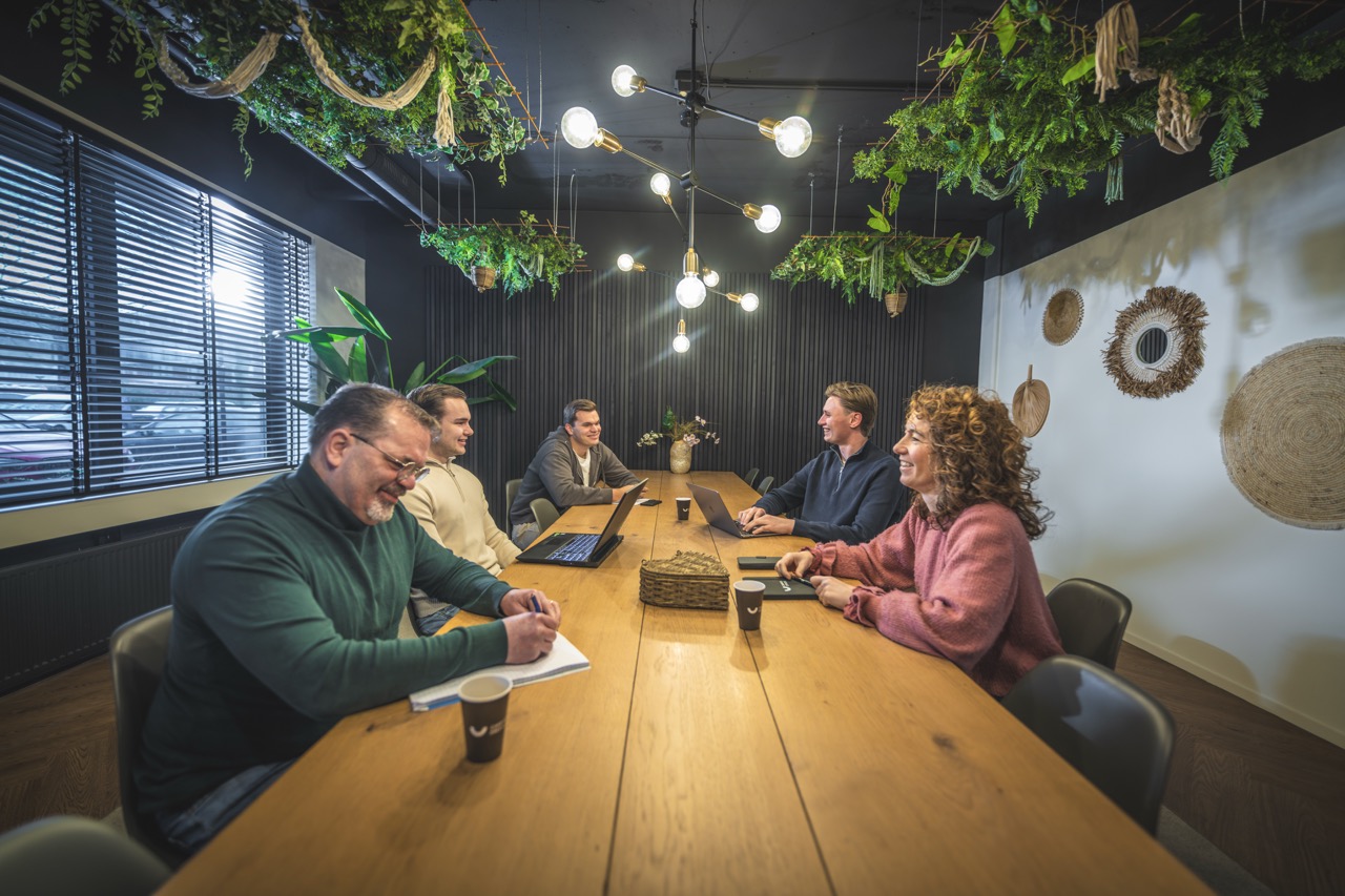 Zes collega’s zitten lachend aan een houten vergadertafel in een modern kantoor met hangende planten en sfeerverlichting.