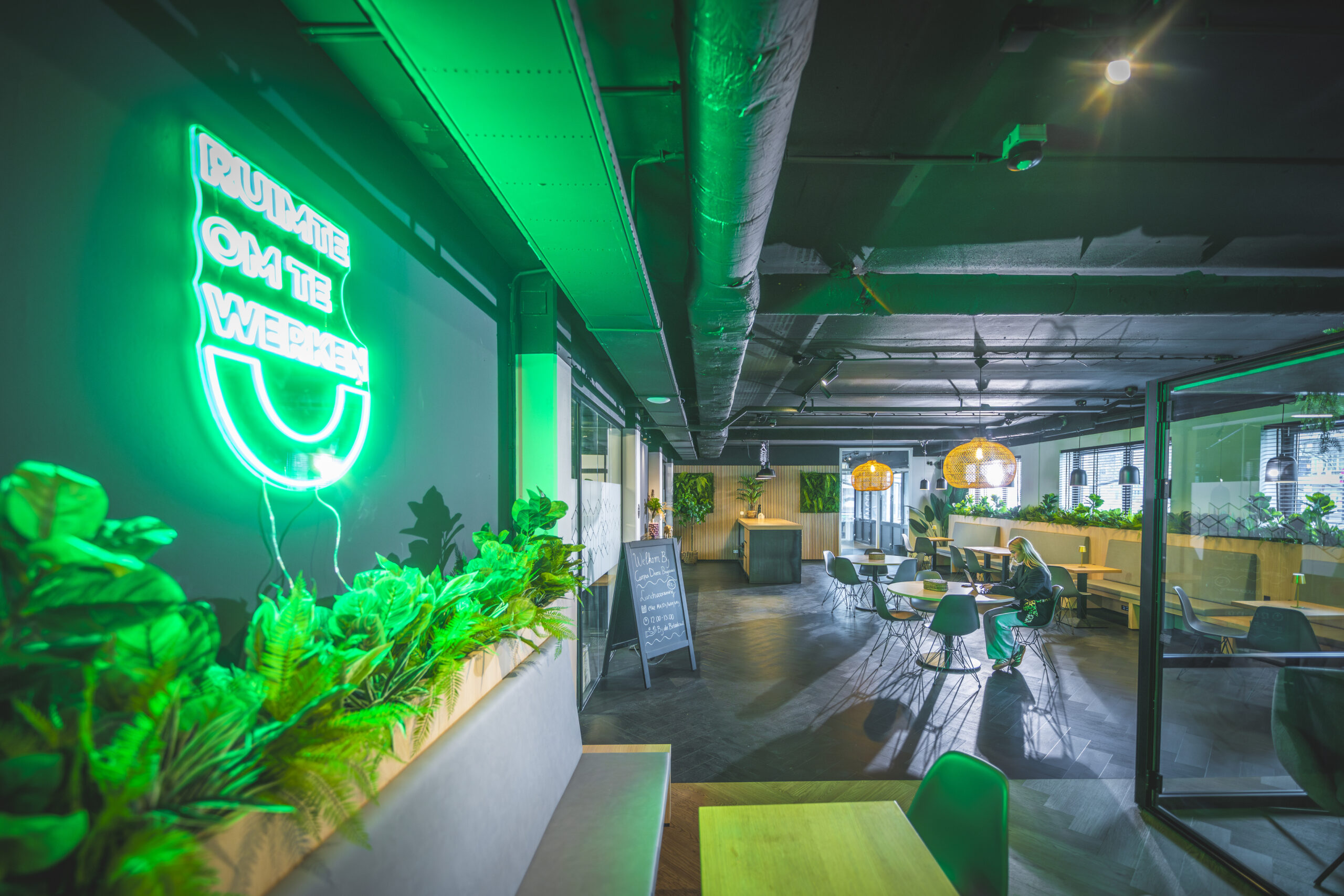 Moderne en groene werkruimte met neonbord "Ruimte om te werken" en een vrouw die aan een tafel werkt.
