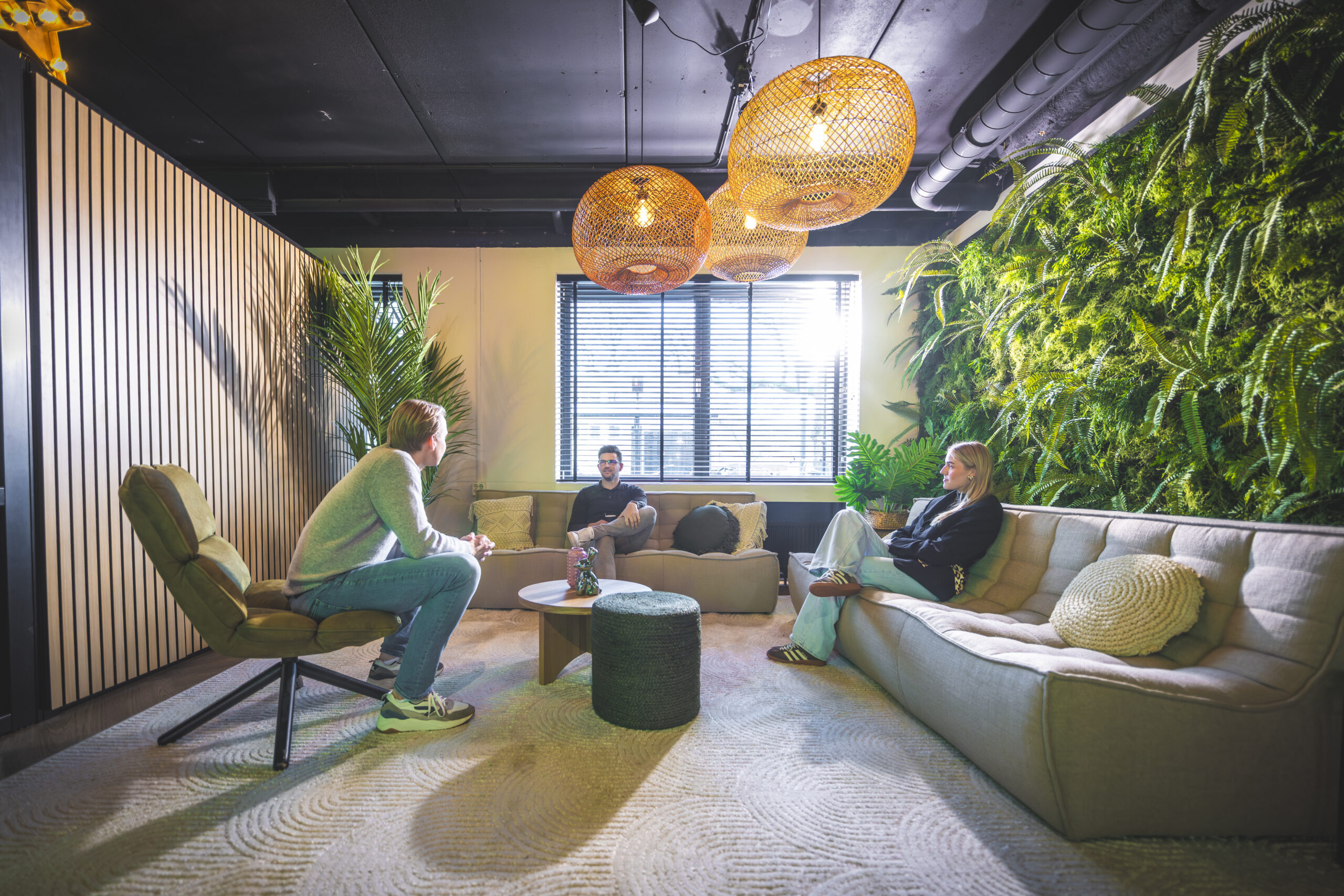 Drie mensen zitten in een modern ingerichte lounge met plantenwand en warme verlichting.