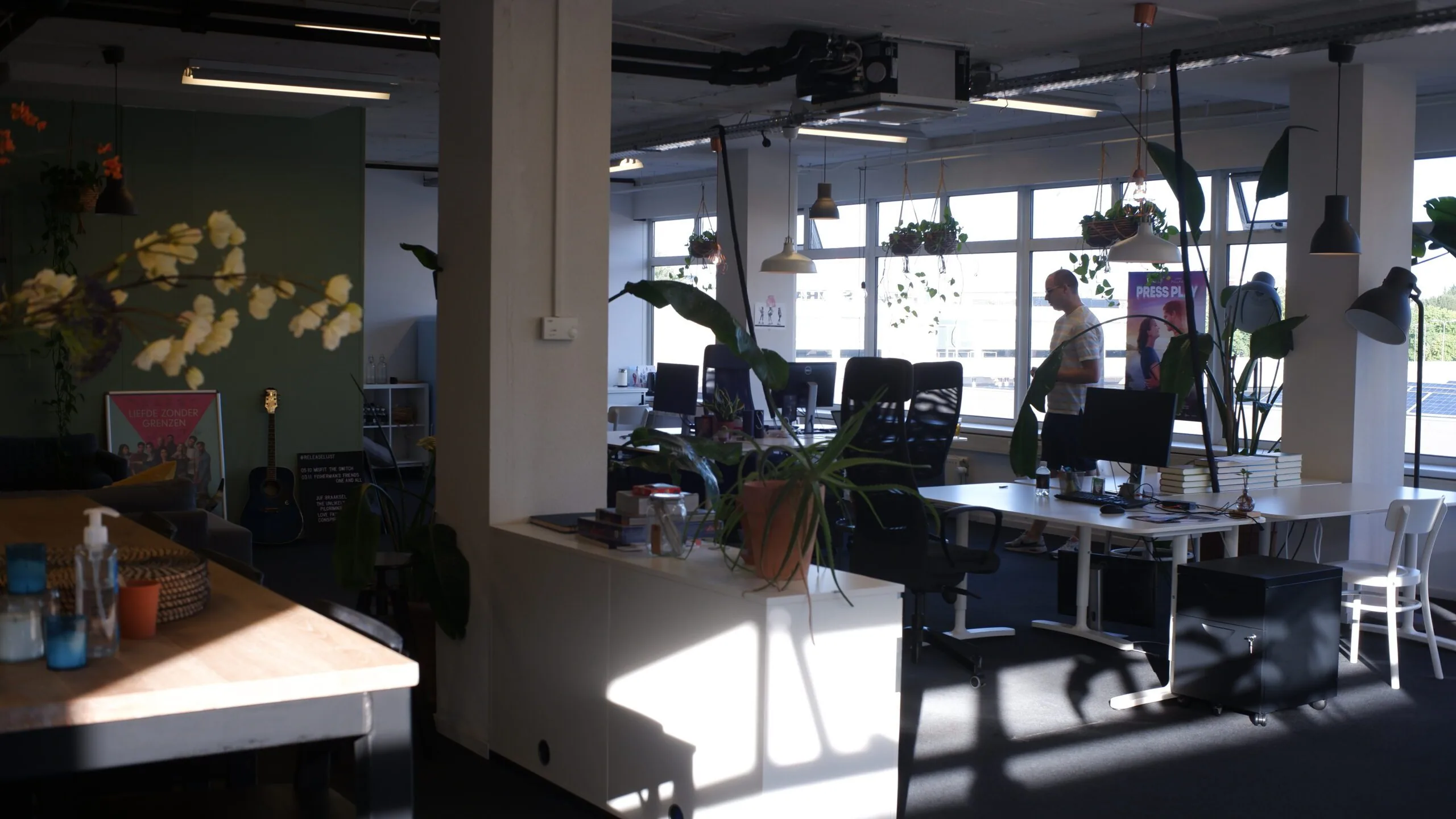 Moderne kantoorruimte met bureaus, planten, hanglampen en een persoon bij het raam.