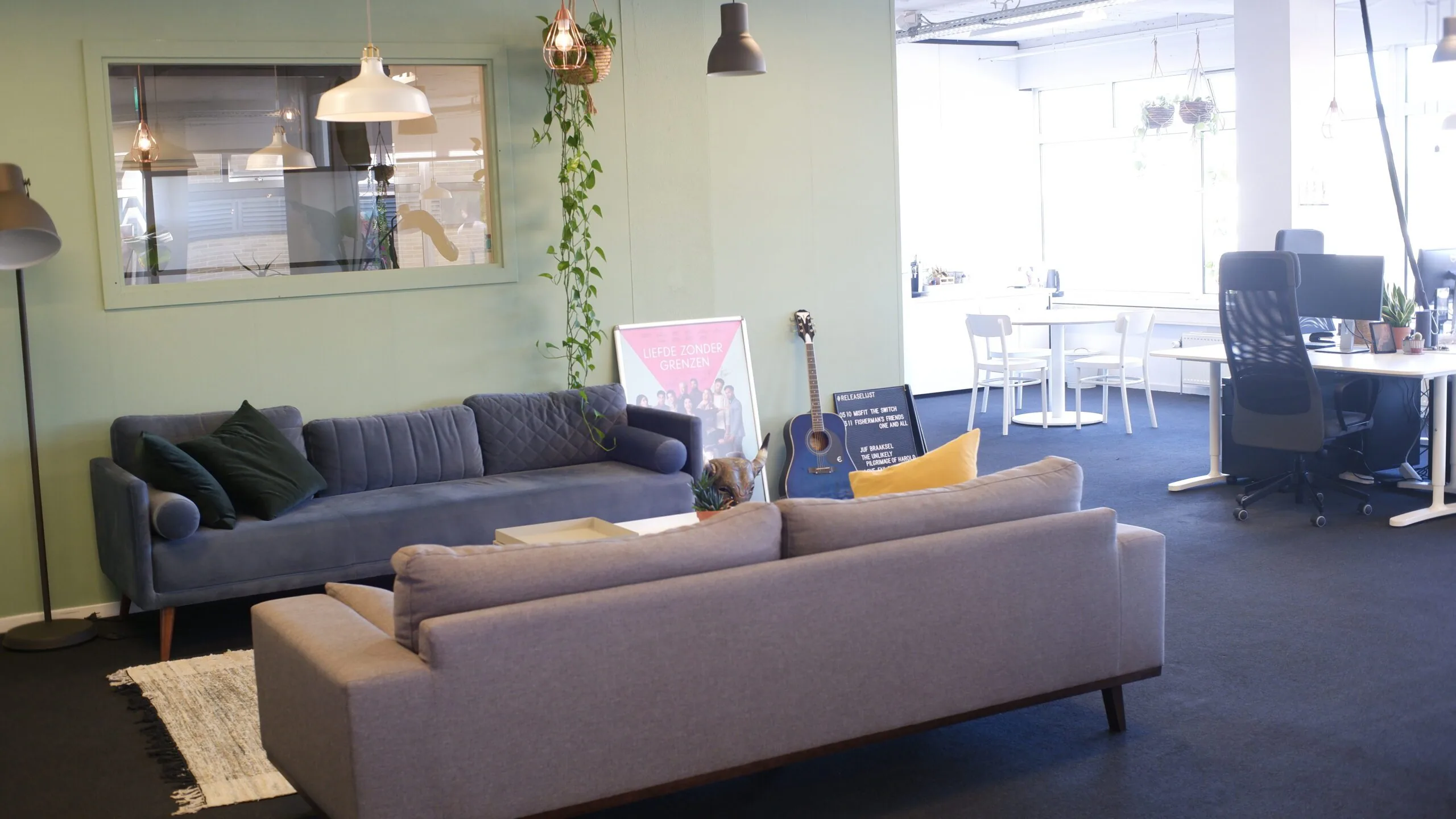 Moderne kantoorruimte met zithoek, planten, een gitaar en werkplekken bij het raam.