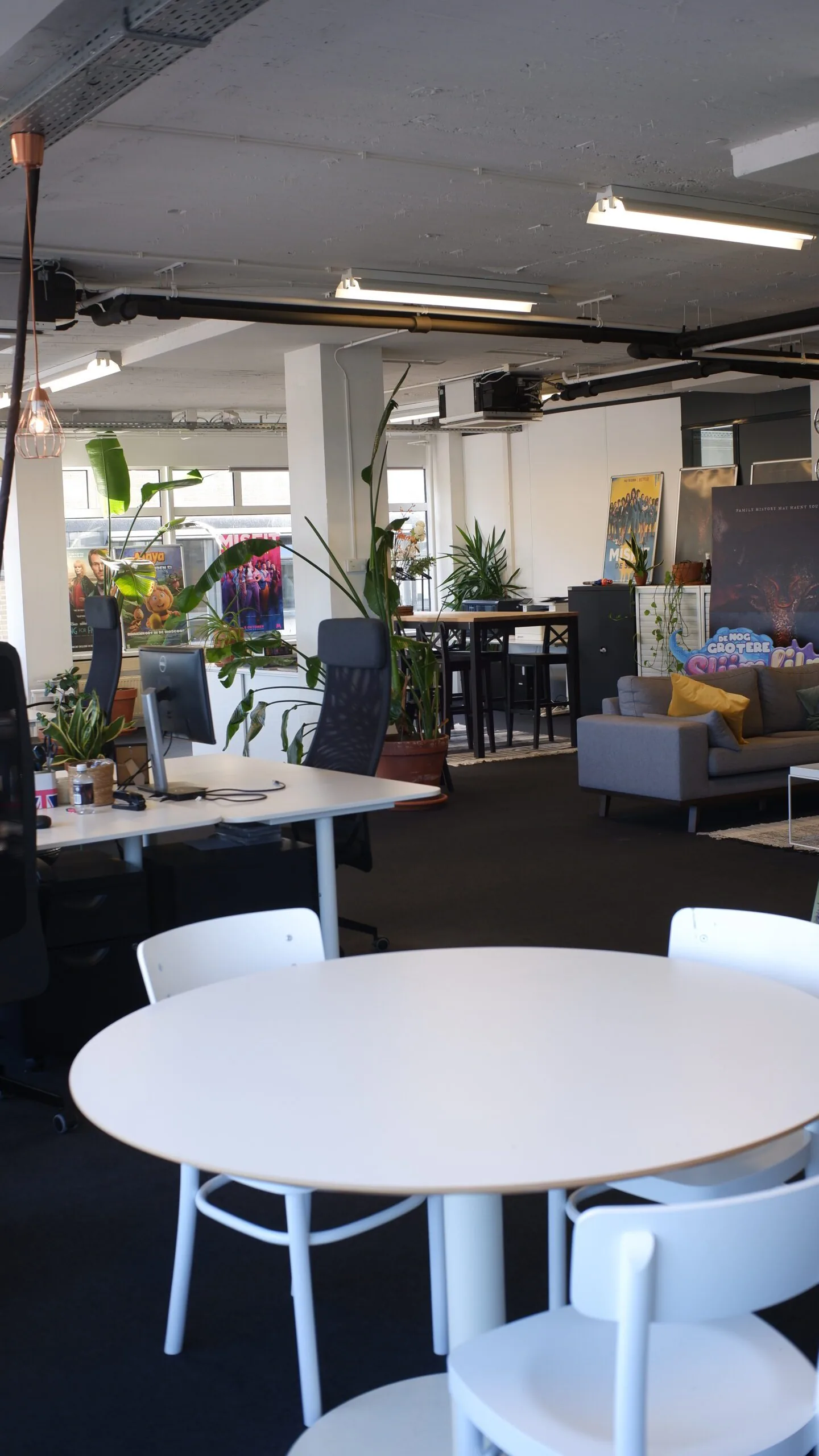 Modern kantoorinterieur met werkplekken, planten, een zithoek en kleurrijke filmposters op de achtergrond.