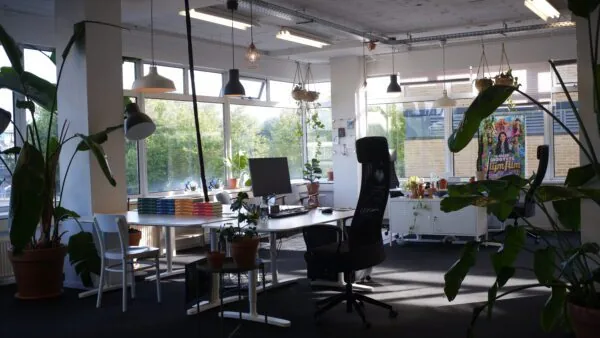 Modern en licht kantoor met veel planten, bureaus, computerschermen en een kleurrijke filmposter op de achtergrond.