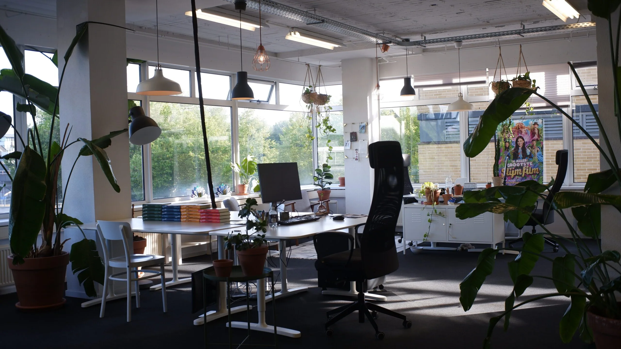 Modern en licht kantoor met veel planten, bureaus, computerschermen en een kleurrijke filmposter op de achtergrond.