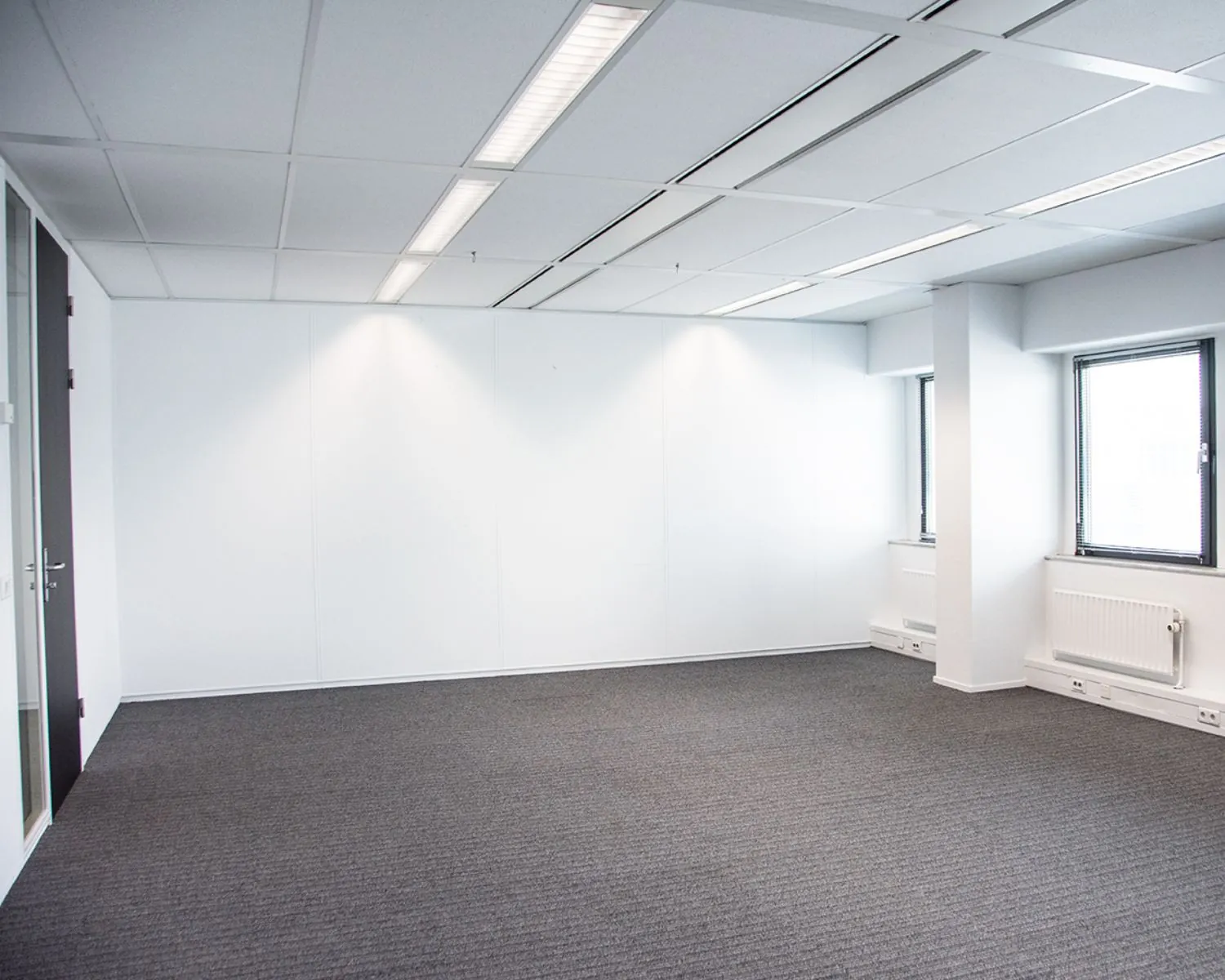 Leeg, licht kantoor met grijze vloerbedekking, witte muren en twee ramen aan de rechterkant.