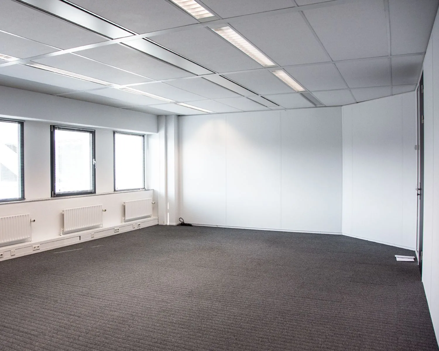 Lege kantoorruimte met grijze tapijtvloer, witte wanden, systeemplafond en ramen met jaloezieën.