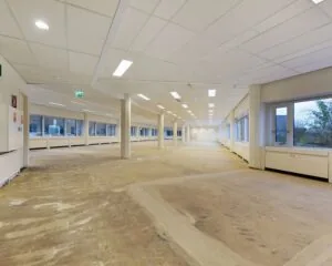 Leegstaand kantoorpand met systeemplafond, grote ramen en kale betonnen vloer.