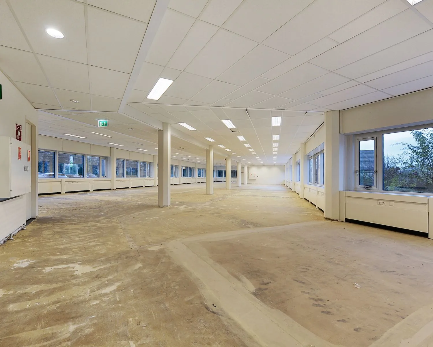 Leegstaand kantoorpand met systeemplafond, grote ramen en kale betonnen vloer.