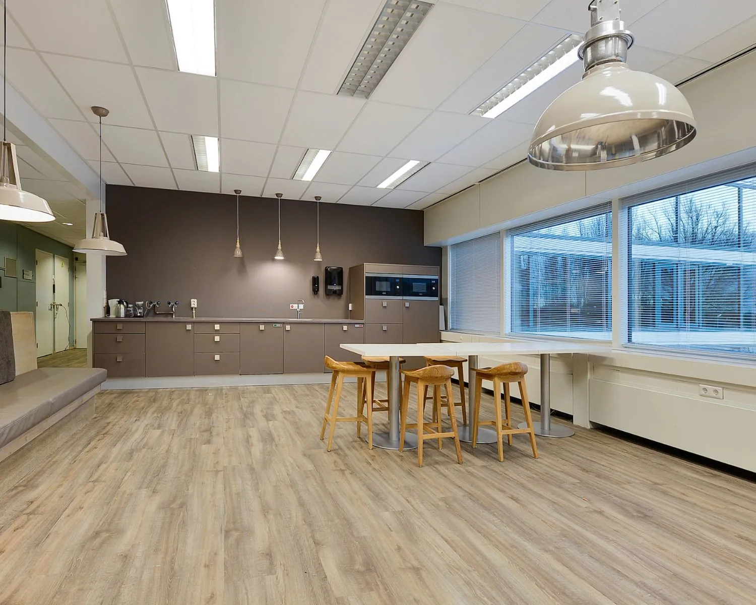 Moderne kantoorkeuken met houten vloer, hoge tafels en barkrukken, en grote ramen met jaloezieën.