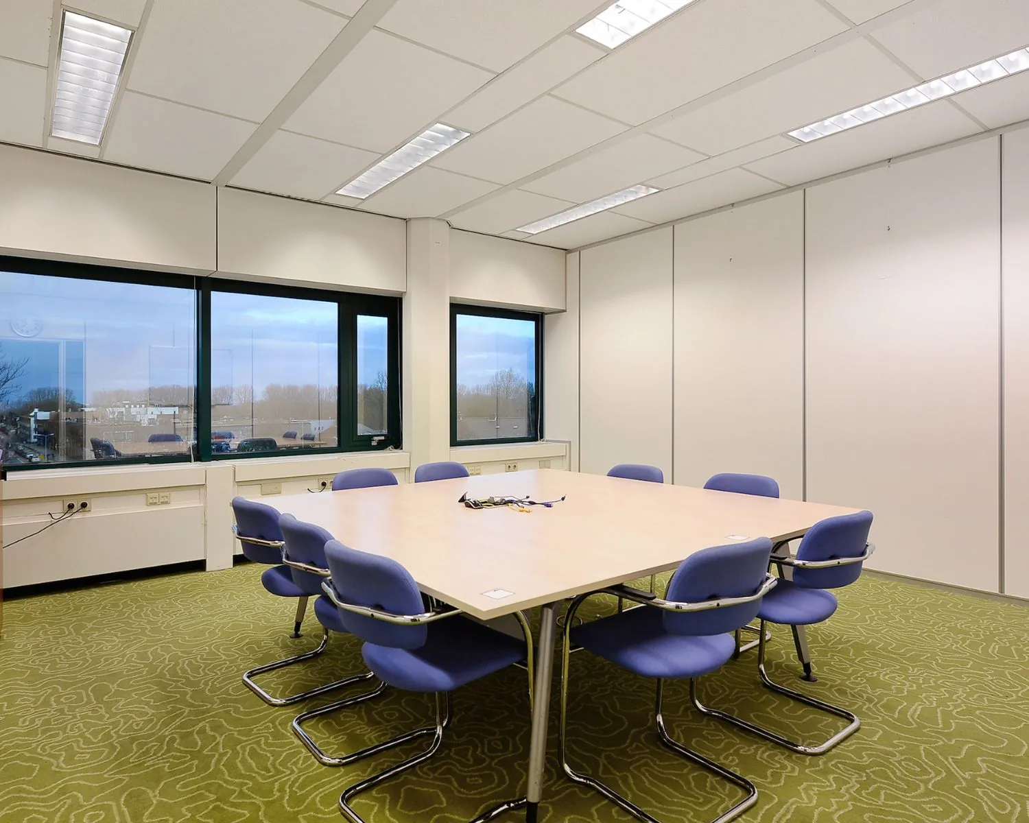 Vergaderruimte met een grote tafel, acht paarse stoelen en uitzicht op een parkeerterrein door drie ramen.