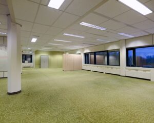 Lege kantooruimte met groen tapijt, systeemplafond en grote ramen aan de rechterzijde.