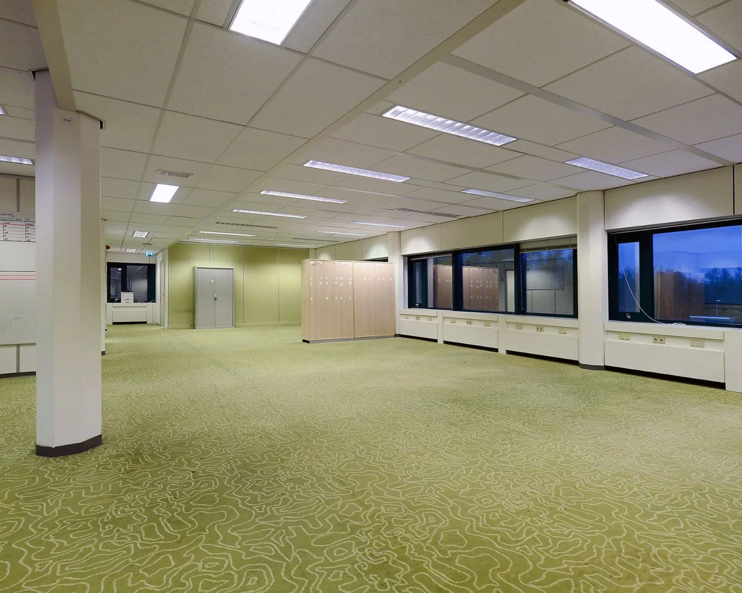 Lege kantooruimte met groen tapijt, systeemplafond en grote ramen aan de rechterzijde.