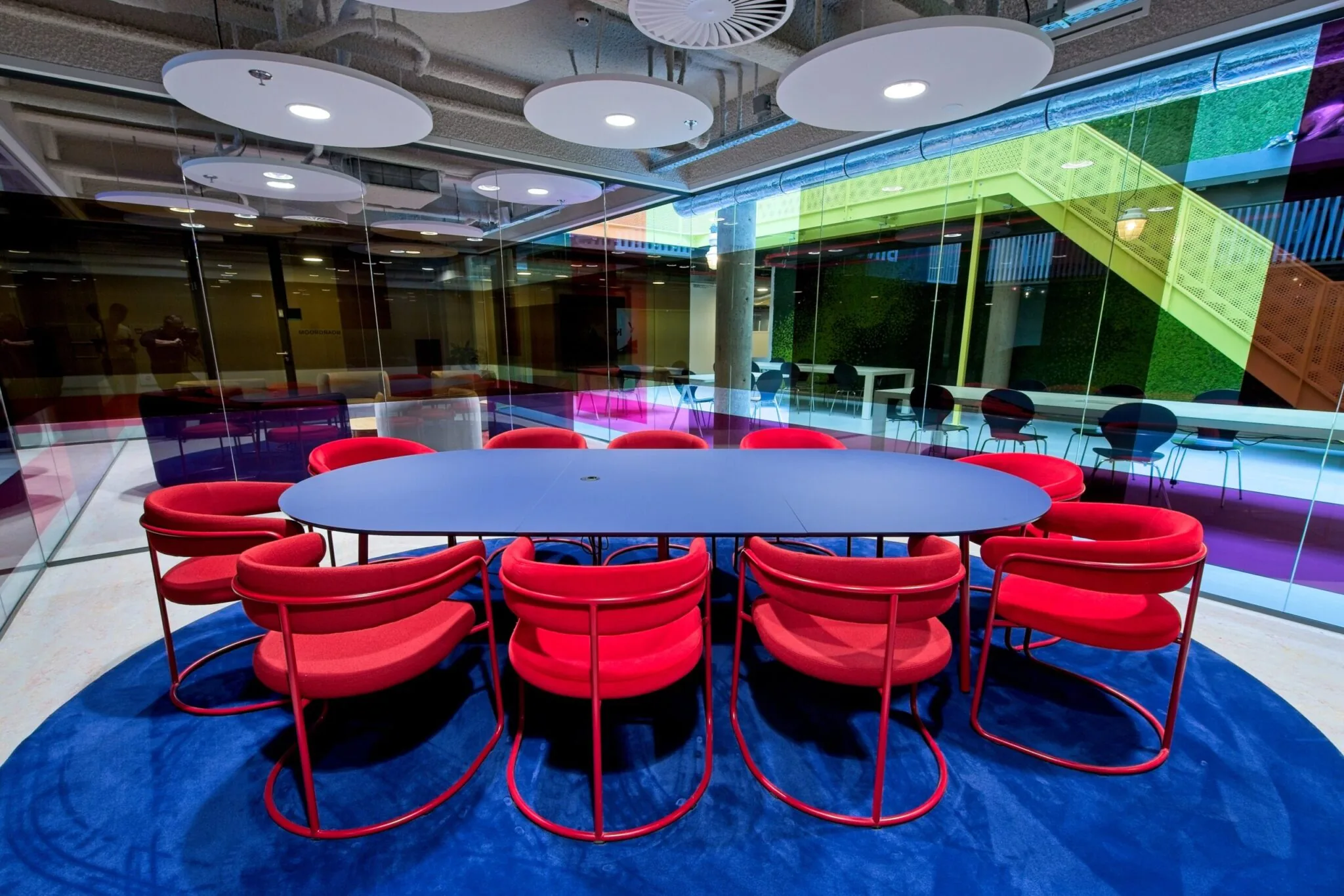 Vergaderruimte met een ovale blauwe tafel en rode stoelen op een blauw tapijt, omringd door glazen wanden.