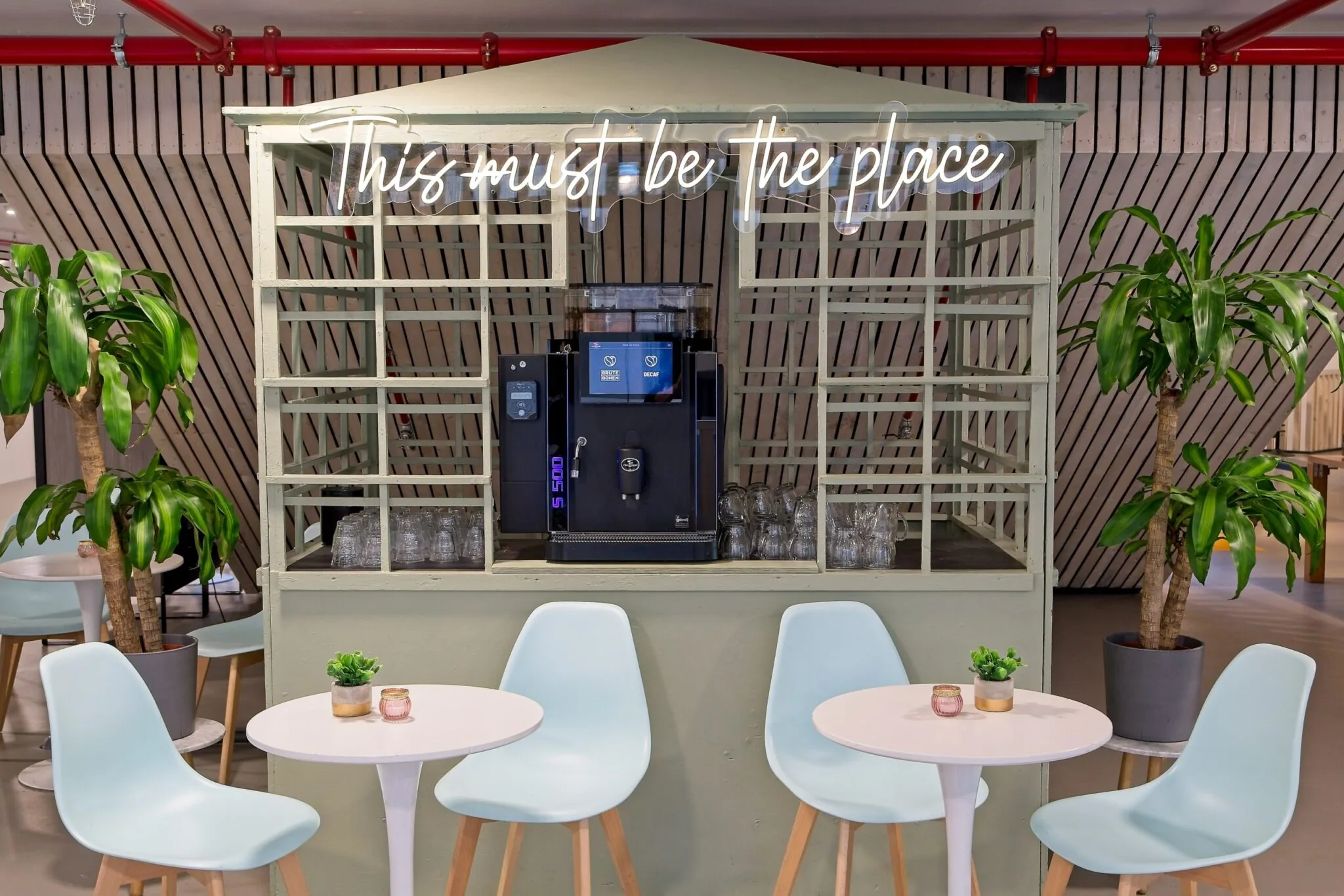 Koffiecorner met automaat, pastelkleurige stoelen en tafels, planten en een lichtbord met de tekst "This must be the place".