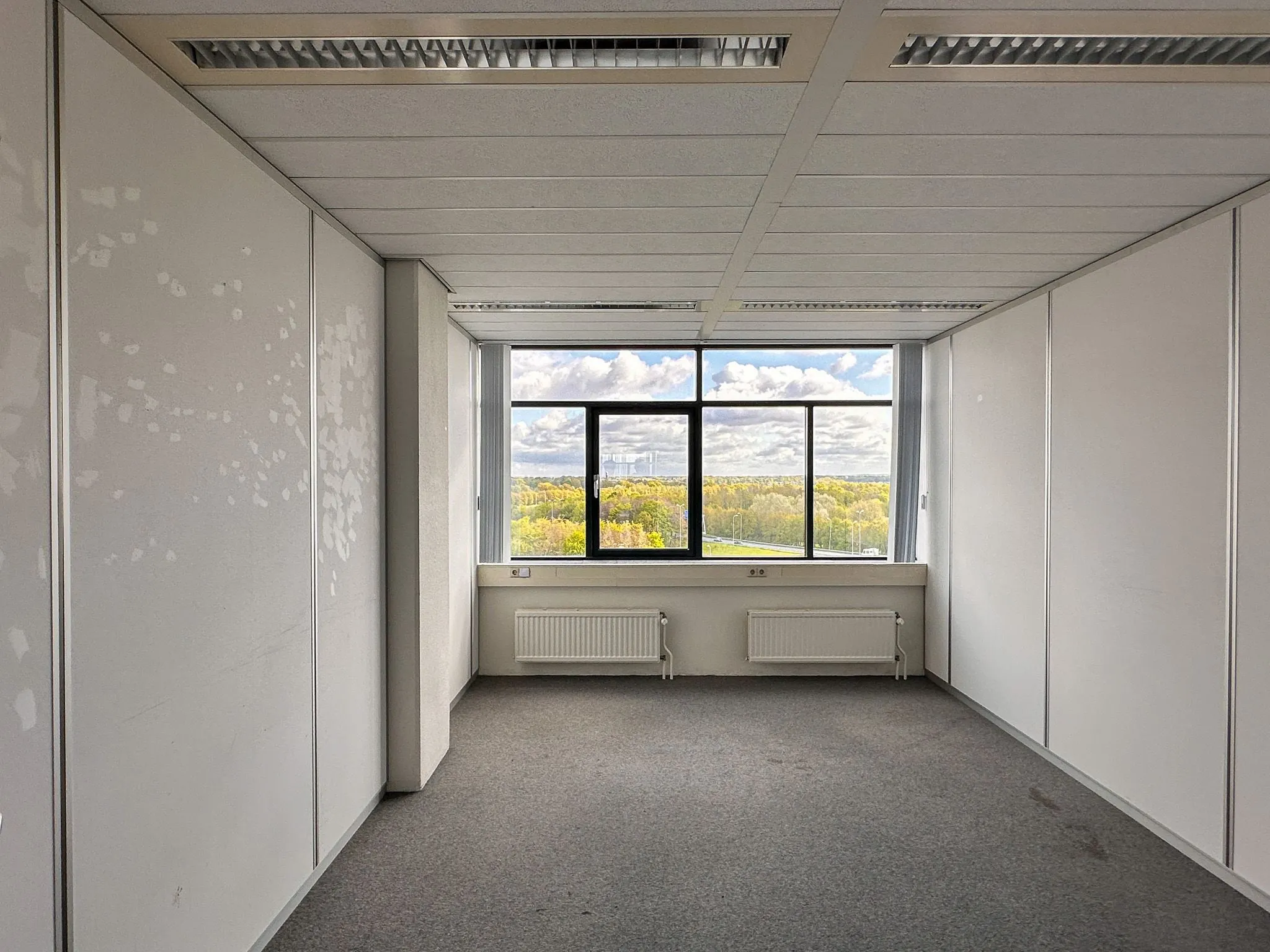 Leeg kantoorruimte op Daltonlaan 200 met groot raam en uitzicht op bomen en wolkenlucht.
