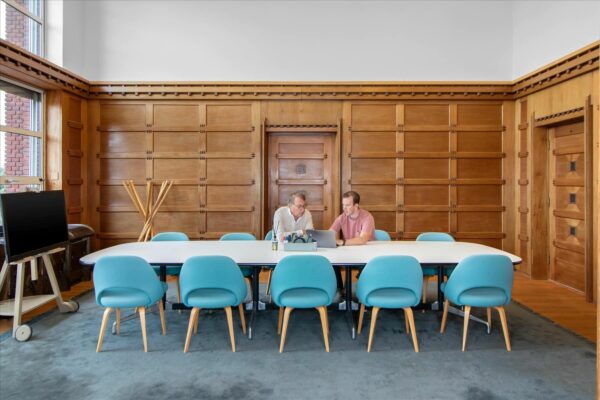 Twee mannen werken samen aan een laptop in een vergaderruimte van De Rode Olifant met houten wandpanelen en turquoise stoelen.