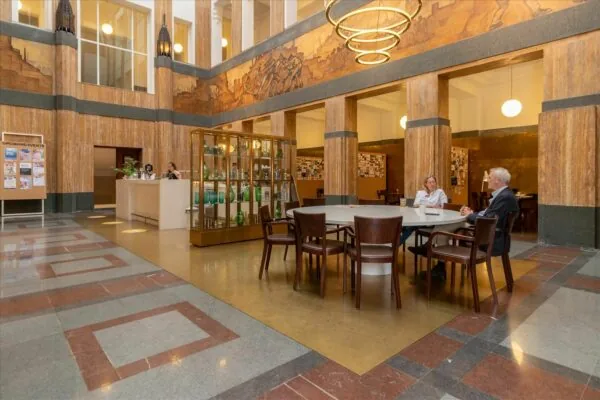 Twee mensen zitten aan een ronde tafel in de kunstzinnige hal van De Rode Olifant met marmeren zuilen en een vitrinekast vol glaswerk.