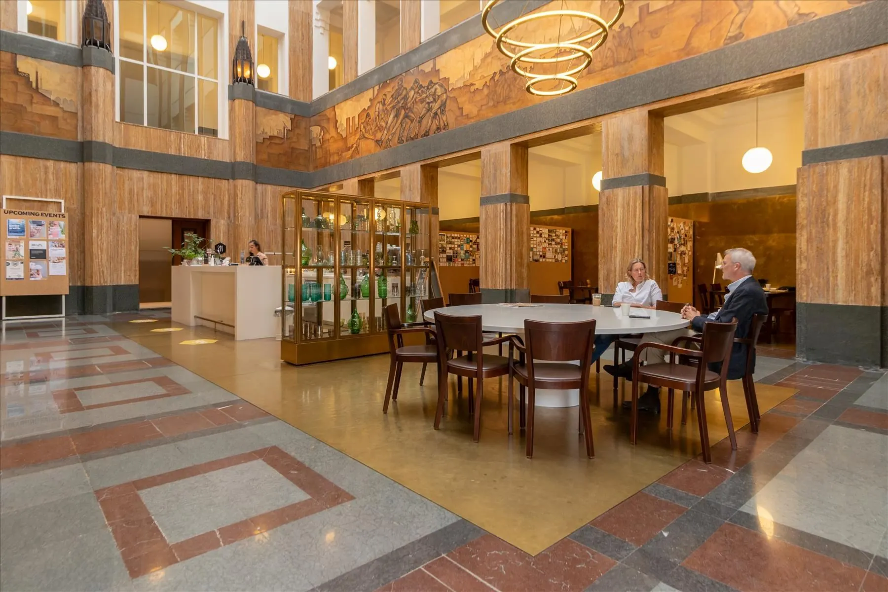 Twee mensen zitten aan een ronde tafel in de kunstzinnige hal van De Rode Olifant met marmeren zuilen en een vitrinekast vol glaswerk.