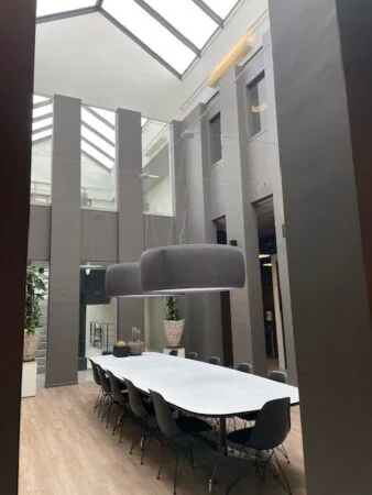 Doorkijk vanuit de keuken naar een modern atrium met een lange vergadertafel en grote hanglampen onder een glazen dak.