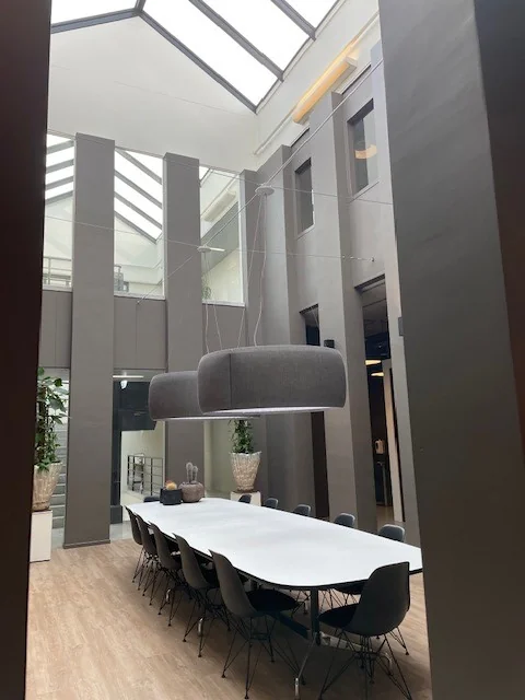 Doorkijk vanuit de keuken naar een modern atrium met een lange vergadertafel en grote hanglampen onder een glazen dak.