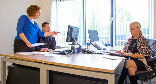 Drie mensen werken samen in een modern kantoor aan de Dr. Nolenslaan 157, met computers en documenten op het bureau.