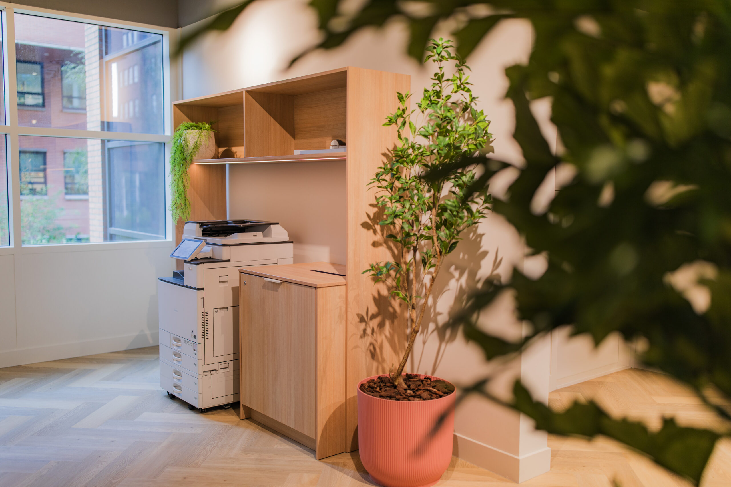 Modern office corner with a printer, wooden shelves, and a potted plant near a large window.