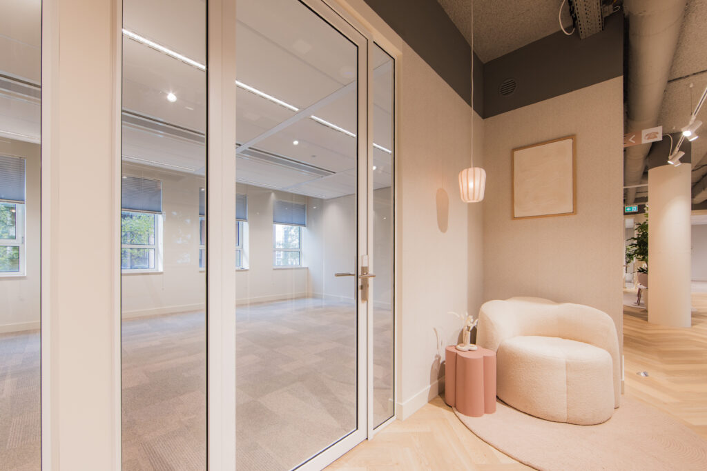 Cozy seating area with a white armchair, pink side table, and pendant light next to a glass door in the Hullenberg building.