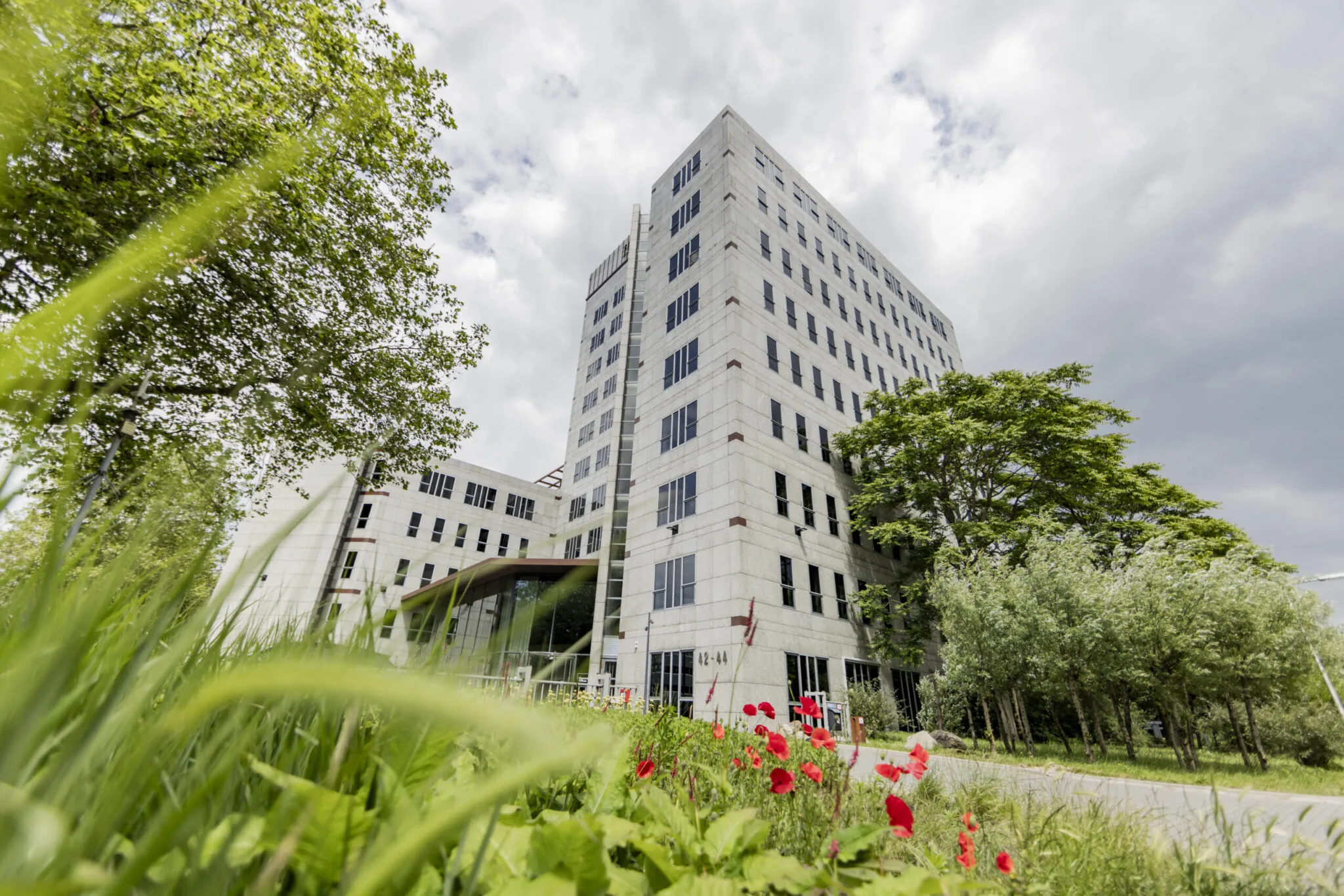 Een modern kantoorgebouw omringd door groen en rode bloemen onder een bewolkte hemel.