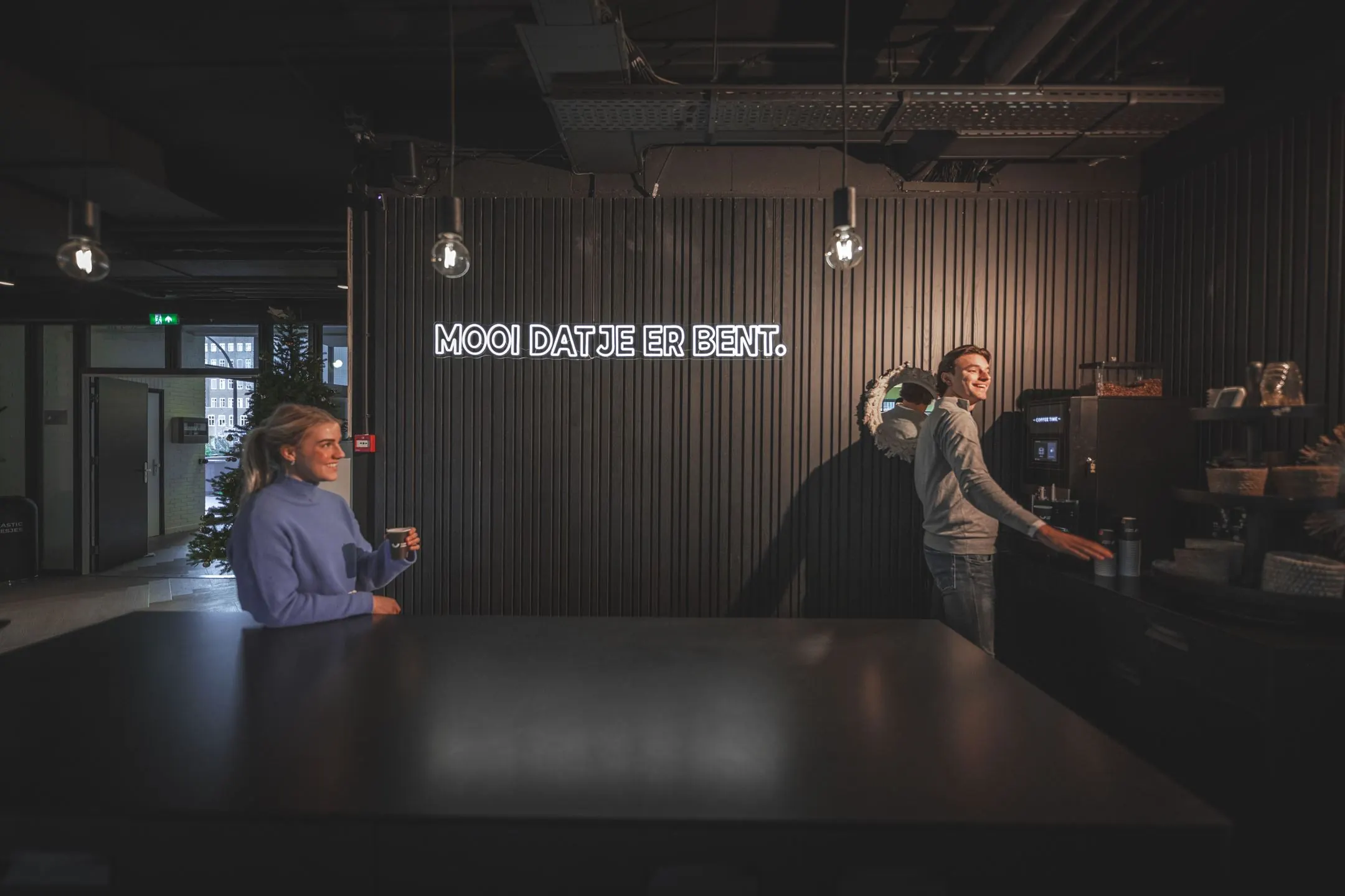 Twee mensen staan bij een koffiemachine in een modern kantoor aan de Egbert Gorterstraat, met op de muur de tekst “Mooi dat je er bent.”