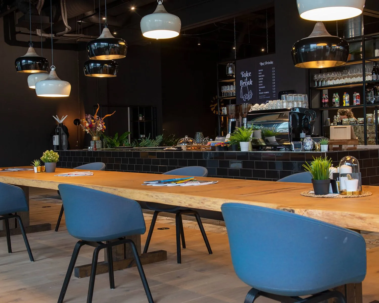 Moderne koffieruimte in Enschede met een lange houten tafel, blauwe stoelen en een stijlvolle koffiebar op de achtergrond.
