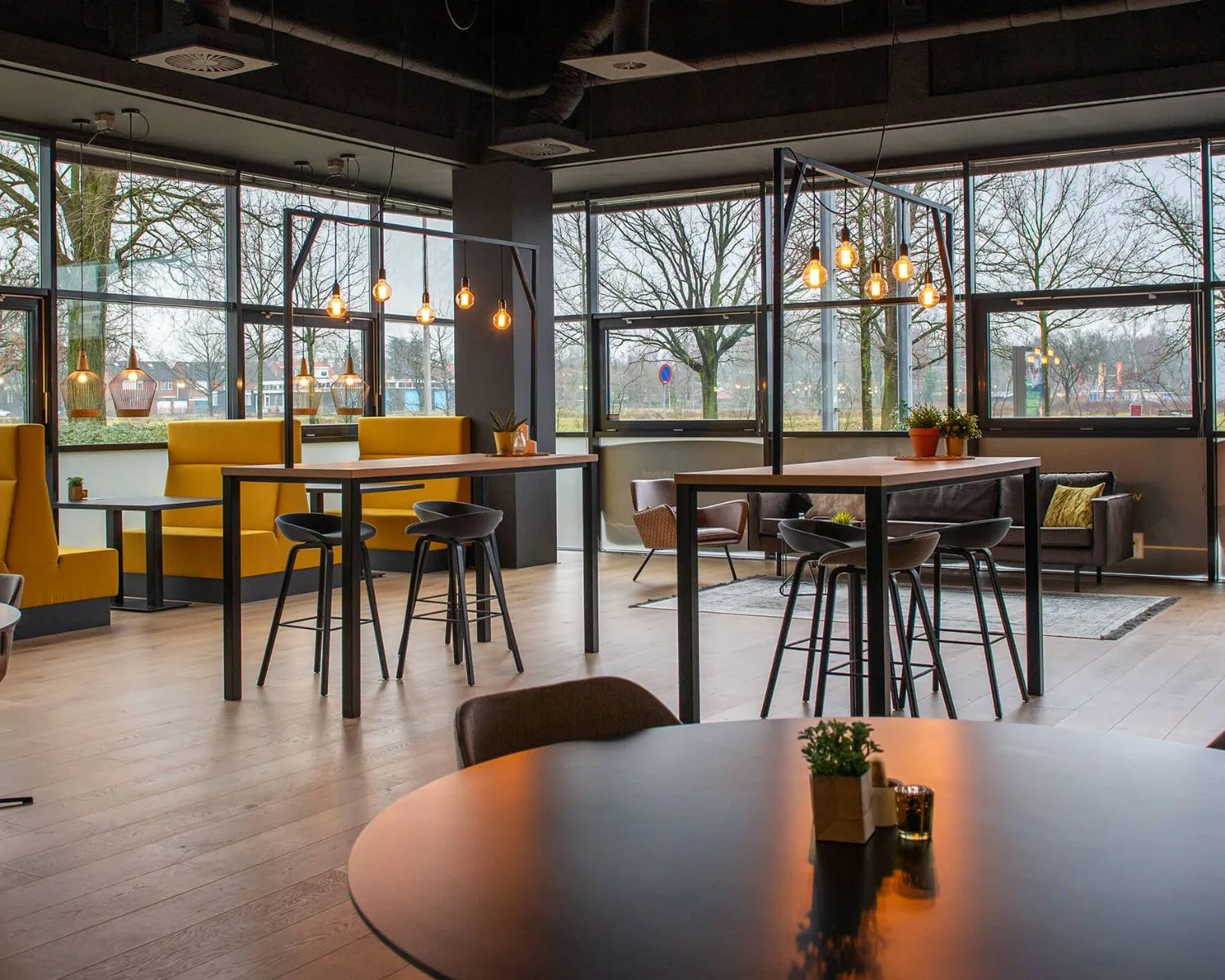 Modern ingerichte horeca- of kantoorlounge in Enschede met hoge tafels, gele zitbanken en grote ramen met uitzicht op bomen.