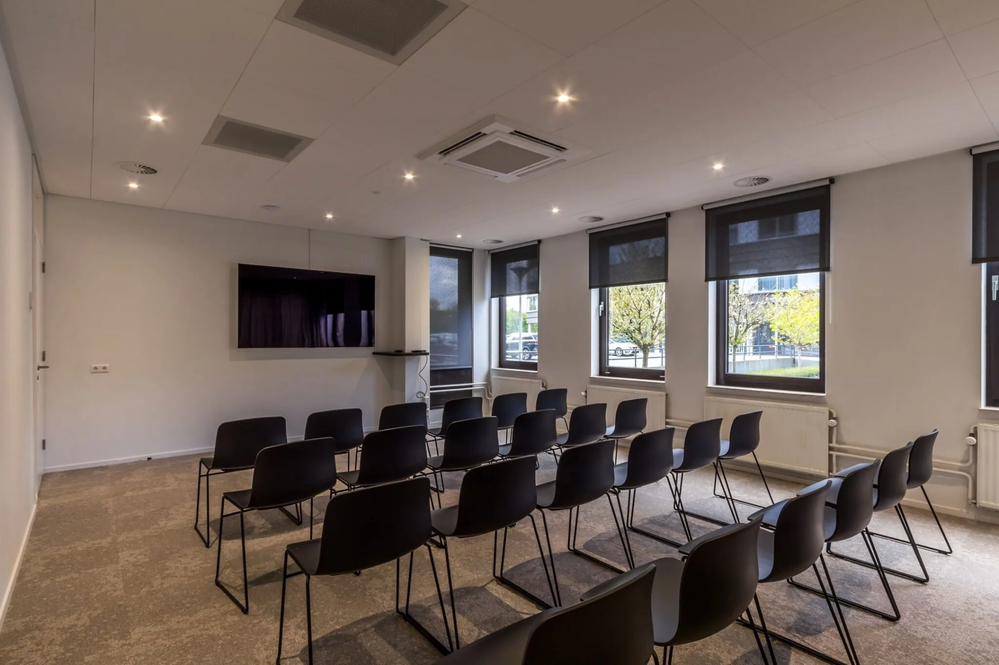 Vergaderruimte op Euclideslaan 265 met stoelen in theateropstelling, een scherm en ramen met rolgordijnen.