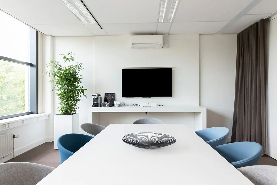 Moderne vergaderruimte aan de Europalaan 16 met witte tafel, blauwe stoelen, flatscreen tv en koffiecorner.