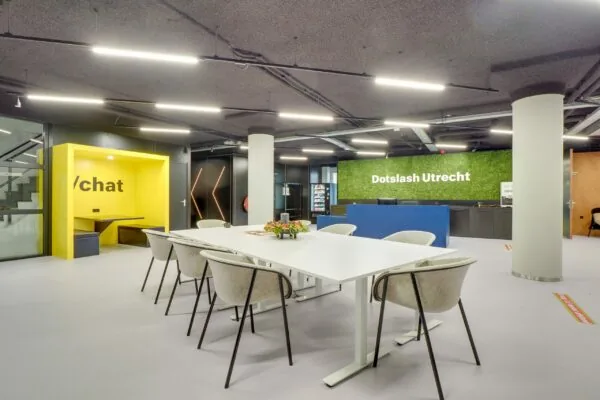 Moderne en kleurrijke kantoorlobby van Dotslash Utrecht aan de Europalaan 400 met een vergadertafel, gele nis met “/chat” en receptiebalie.