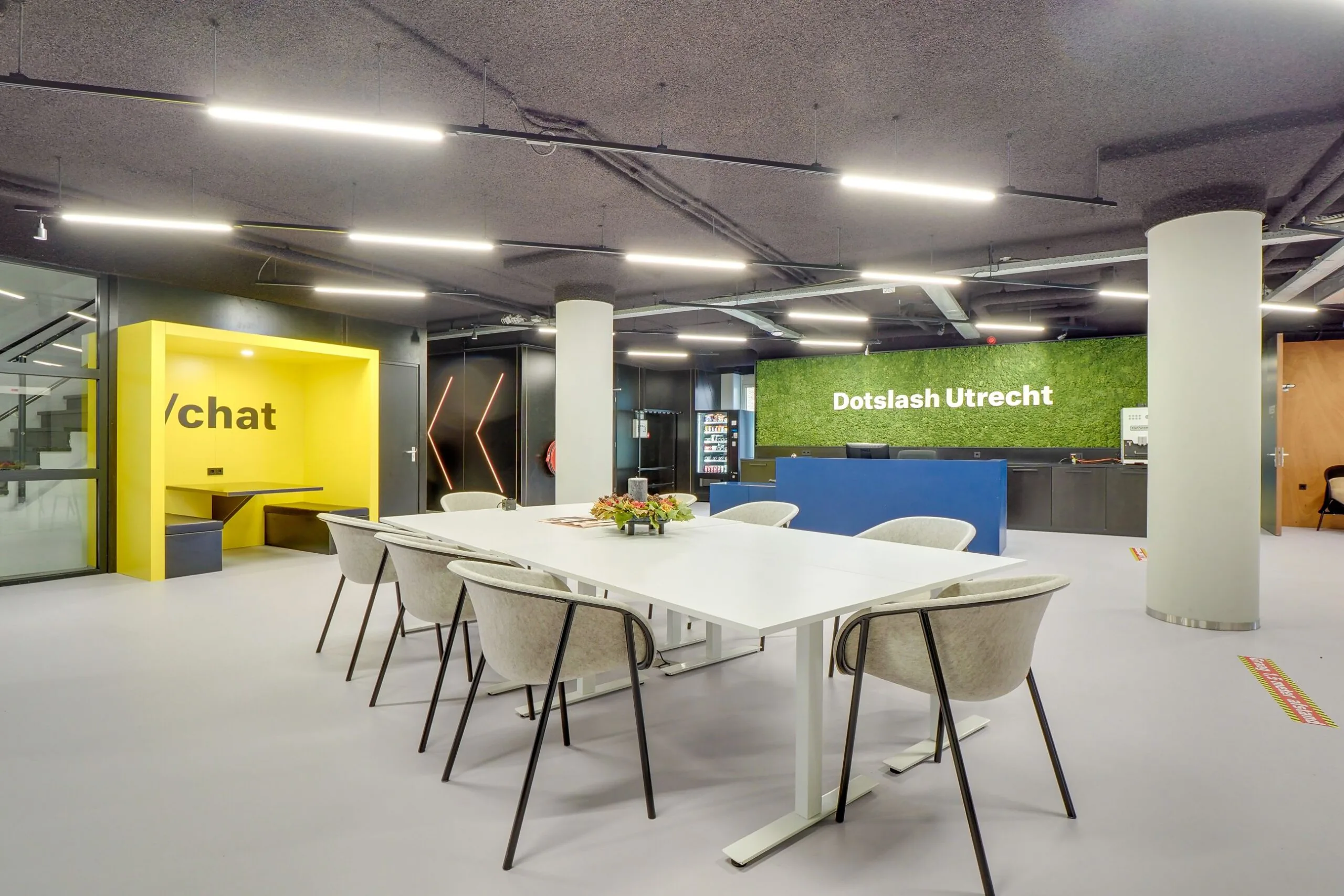 Moderne en kleurrijke kantoorlobby van Dotslash Utrecht aan de Europalaan 400 met een vergadertafel, gele nis met “/chat” en receptiebalie.