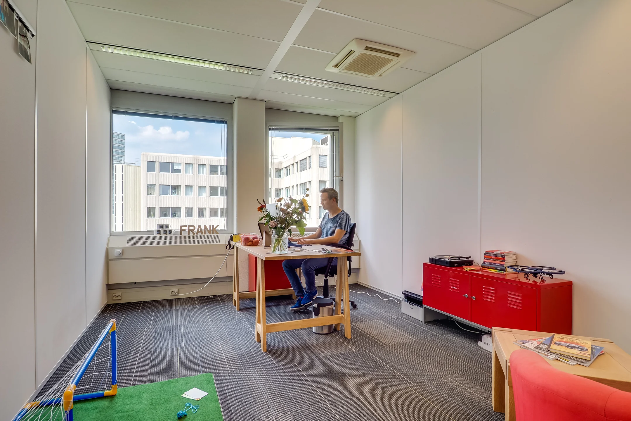 Man werkt op een kantoor aan een bureau bij het raam met uitzicht op een gebouw met het woord "FRANK".