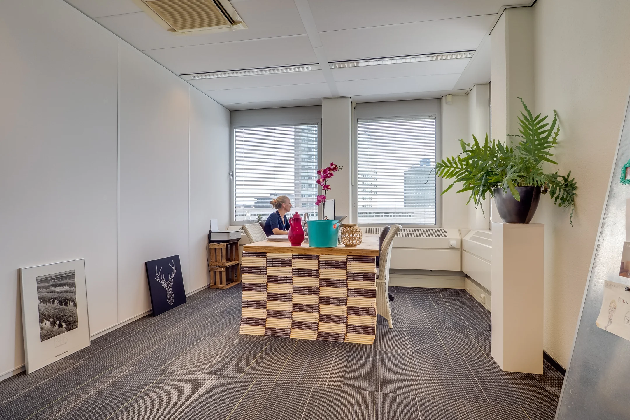 Modern kantoor met een houten bureau, planten en een vrouw die uit het raam kijkt.