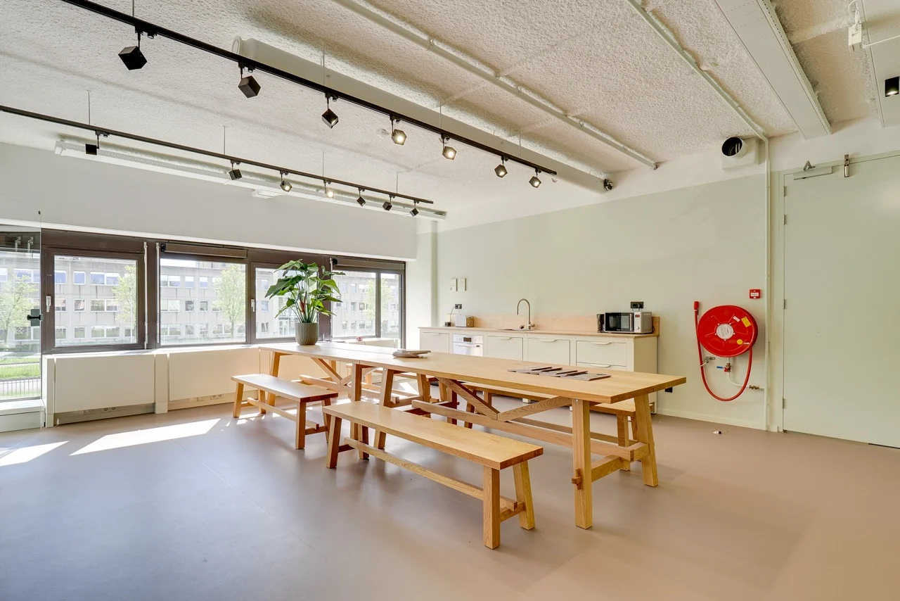 Moderne, lichte kantineruimte aan de Europalaan 93 met houten picknicktafels, een keukenblok en grote ramen met uitzicht op een kantoorgebouw.