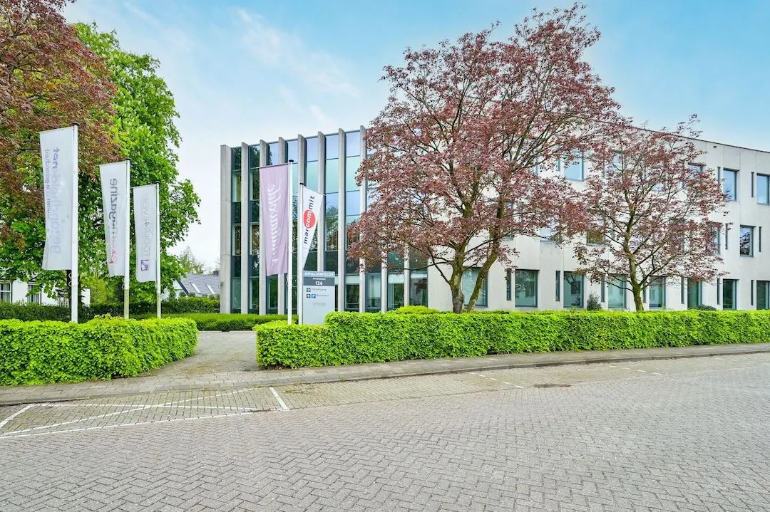 Modern kantoorgebouw FlexOffiZ in Baarn met vlaggen, bomen en een verzorgde groene haag.