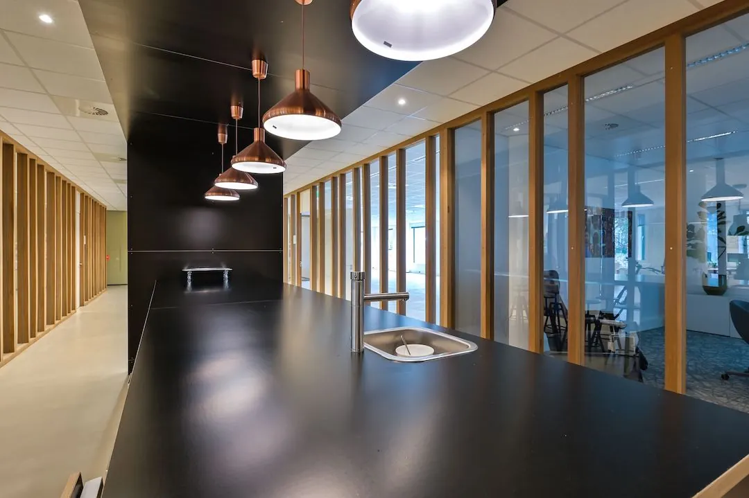 Moderne kitchenette met zwarte bar, koperen hanglampen en glazen wand in FlexOffiZ Baarn.