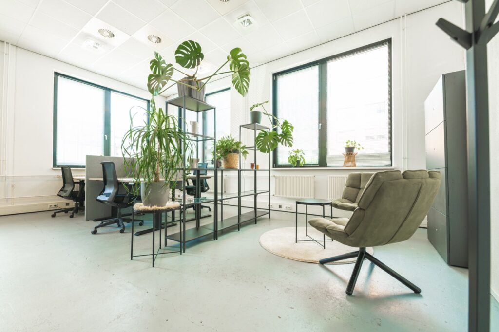 Moderne flexwerkruimte met bureaus, loungezitjes en veel groene planten bij grote ramen.