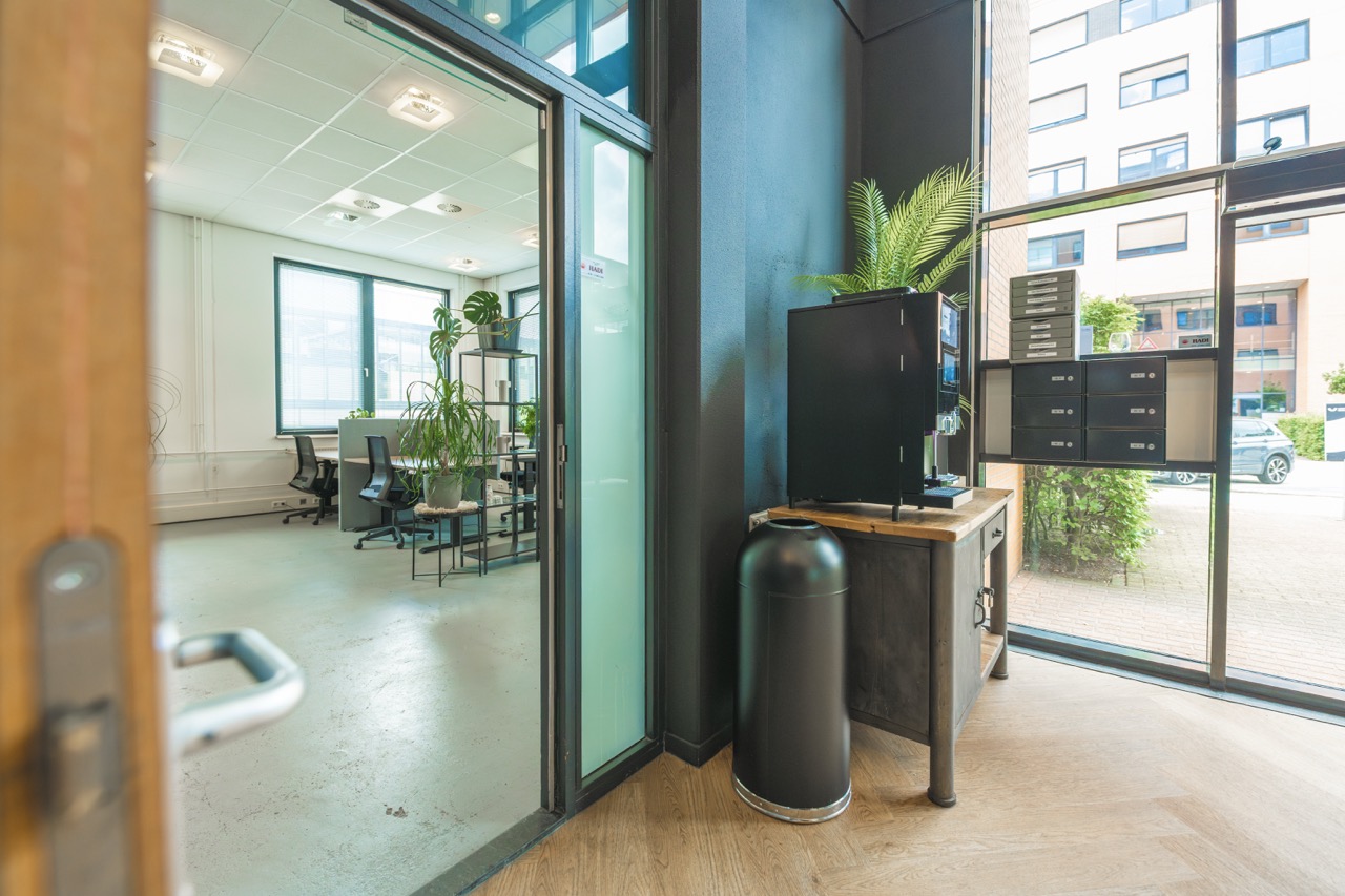 Moderne flexwerkruimte met bureaus, planten en een koffiemachine in een lichte, open kantooromgeving.