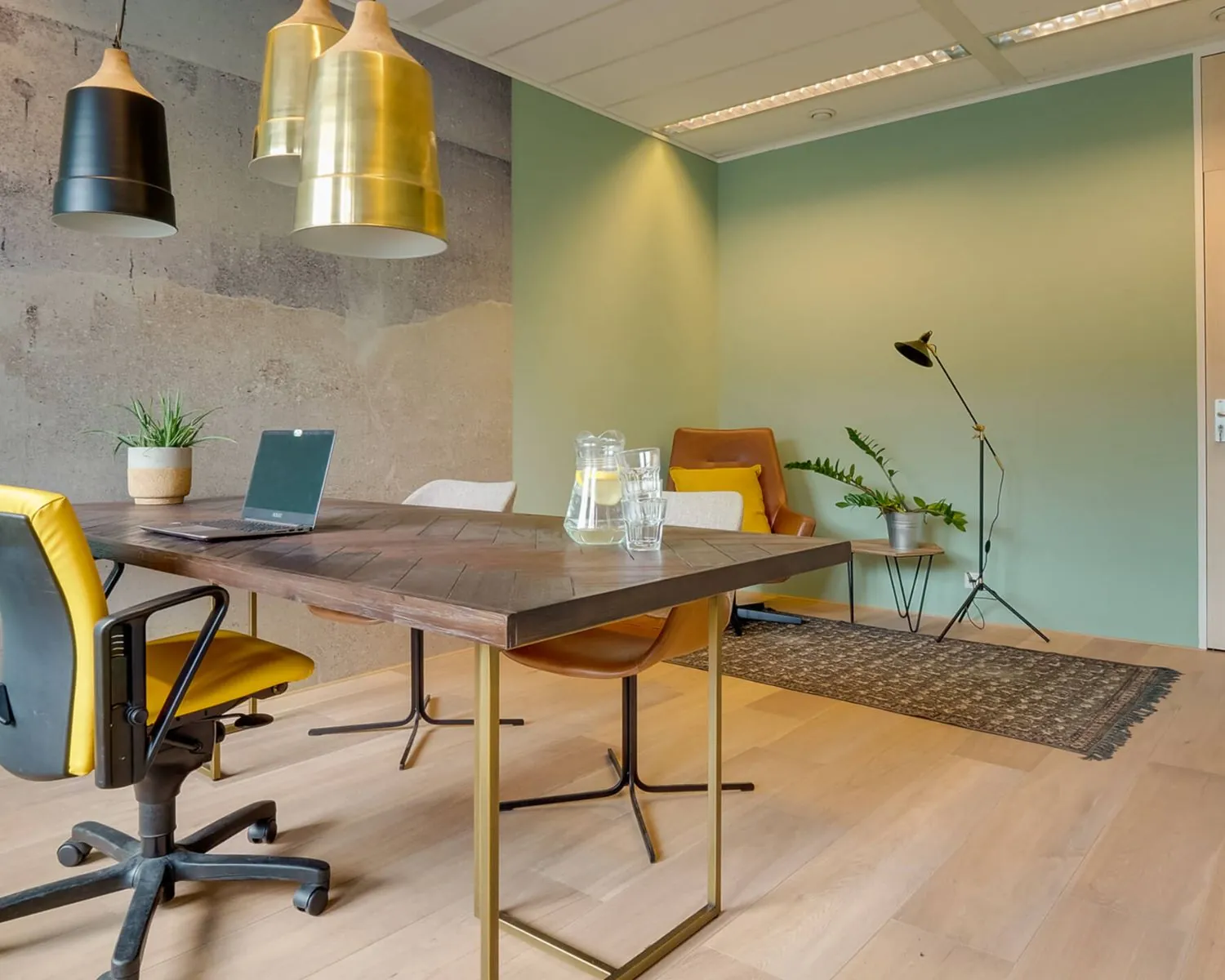 Modern ingericht kantoor met houten tafel, gele accenten, designverlichting en zithoek tegen een groen- en betonlook muur.