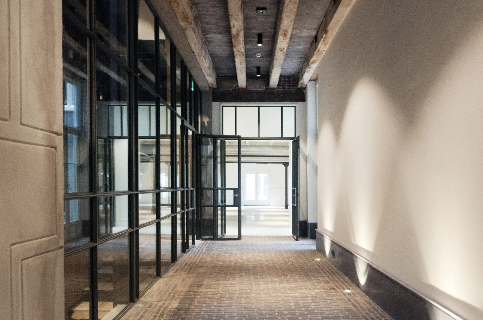 Moderne gang met glazen deuren, bakstenen vloer en zichtbare houten balken aan het plafond.