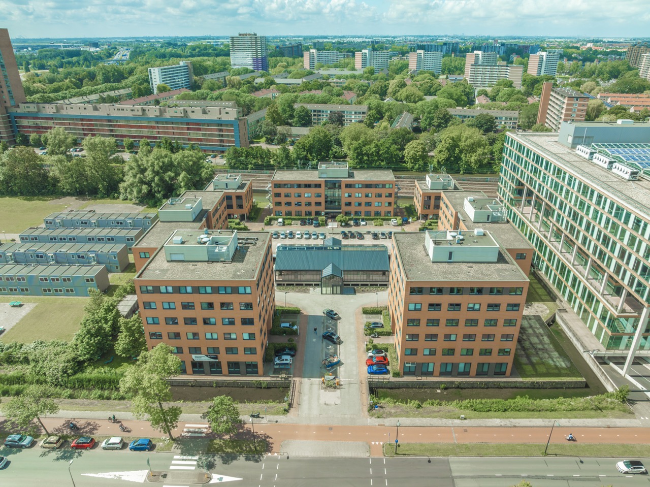 Luchtfoto van een complex met meerdere grote, oranje bakstenen kantoorgebouwen en omliggende hoogbouw in een groene stedelijke omgeving.