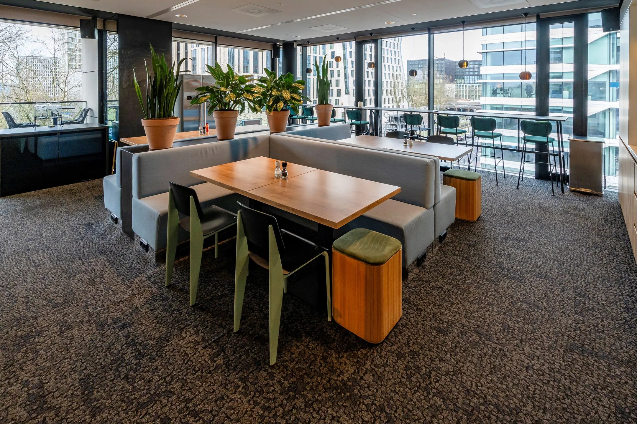Moderne bedrijfsrestaurant op Gustav Mahlerlaan 1025 met houten tafels, grijze banken en grote ramen met uitzicht op kantoorgebouwen.