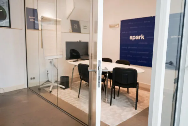 Vergaderruimte met glazen wanden, een ovale tafel, stoelen, een flip-over en een blauw bord met de tekst "spark" aan de muur.