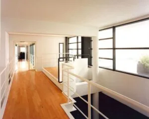 Moderne, lichte hal met houten vloer, witte balustrades en grote ramen aan de rechterzijde.