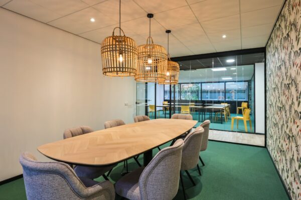 Vergaderruimte aan de Havenweg 24 met een ovale houten tafel, gestoffeerde stoelen en rotan hanglampen, grenzend aan een ruimte met gele stoelen achter een glazen wand.